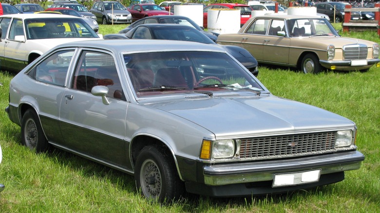 A silver Chevy Citation parked on a grass field with other cars