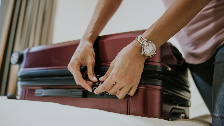 A person wearing a watch locking a maroon suitcase.