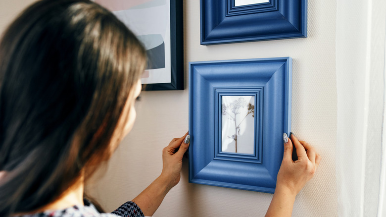 A woman hangs a photo in a blue wooden frame on a cream-colored wall.