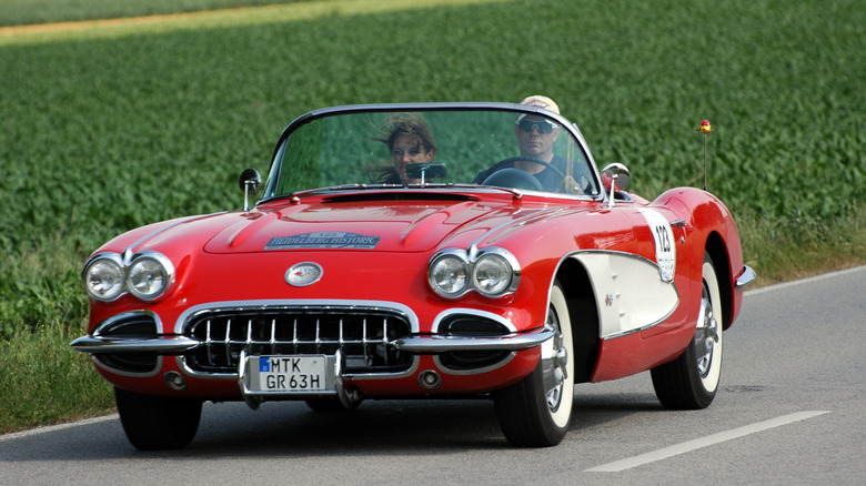 Red 1960 Chevrolet Corvette on country road, male driver with white baseball cap and female passenger with windblown hair