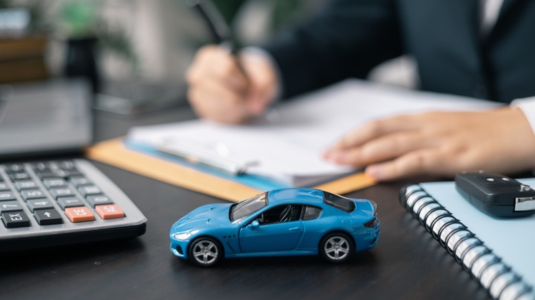 A dealership employee writing notes on a desk as a miniature toy car is perched on the front of the table, along with a calculator, car key, and a journal