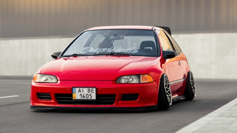 A slammed red car, low to the ground and with wheels slanted outward, and with the words Drop Mode in cursive on its windshield