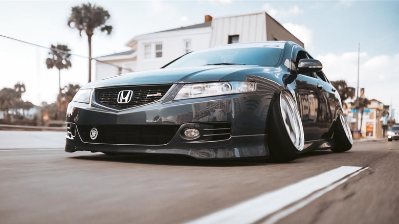 A slammed gray Honda with white rims drives down a street with palm trees in the background