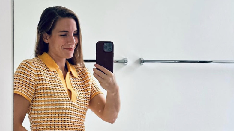 Genet taking a mirror selfie with her iPhone while wearing a yellow and white knitted shirt