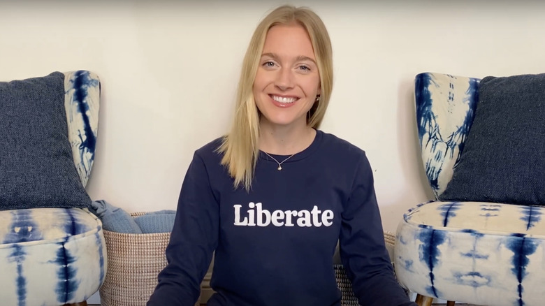 Olivia Bowser wearing a Liberate-branded long-sleeve shirt as she sits in between two blue and white chairs with gray throw pillows and some brown bins at the back