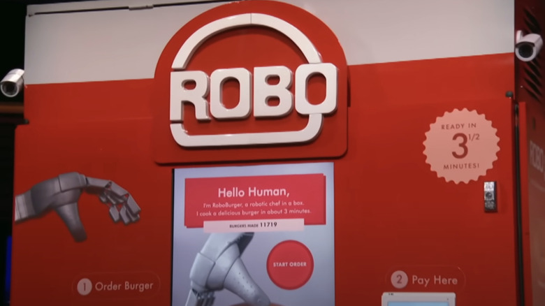 The front of the RoboBurger machine, featuring its logo, an interactive screen, monitoring cameras on the side, and printed instructions on how to order and pay