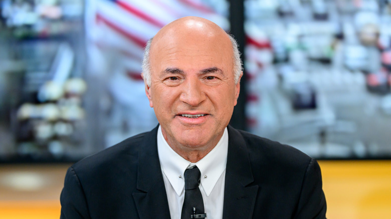 Kevin O'Leary smiling in front of the camera while wearing a suit and tie