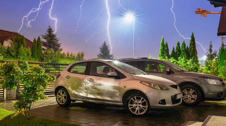 Two cars parked in a driveway with lightning storm in the background