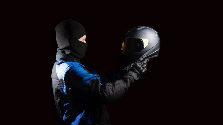 A person wearing a balaclava holding up a gray motorcycle helmet.