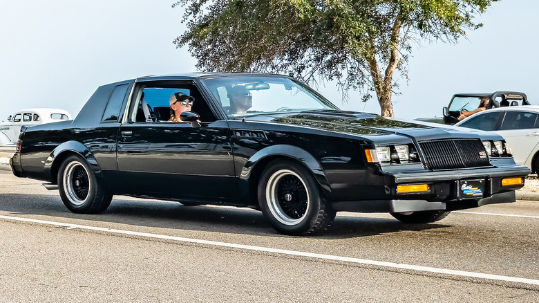 Black Buick GNX driving on a road with a tree and other cars in the background