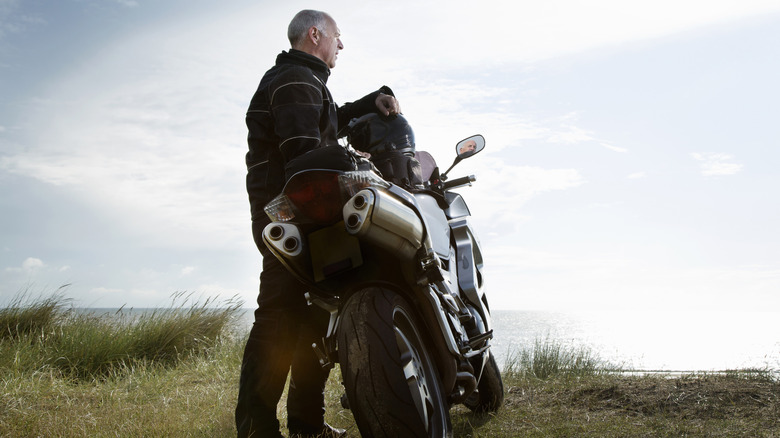 A middle-aged man stands next to a motorcycle in a grassy field