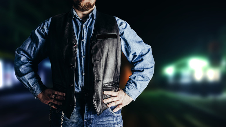 A man in jeans shirt and black biker vest standing in front of a neon night city background