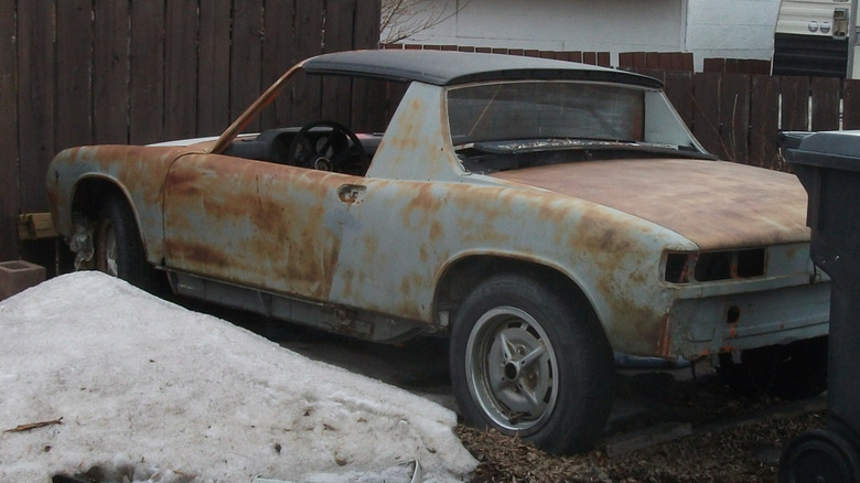 A rusty Porsche 914 parked on a driveway next to dirty snow and a trash can.