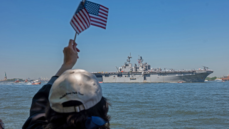 A person wearing a cap raising an American flaglet on a dock as a U.S. Navy ship disembarks