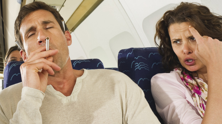 A man smoking a cigarette next to a woman on an airplane