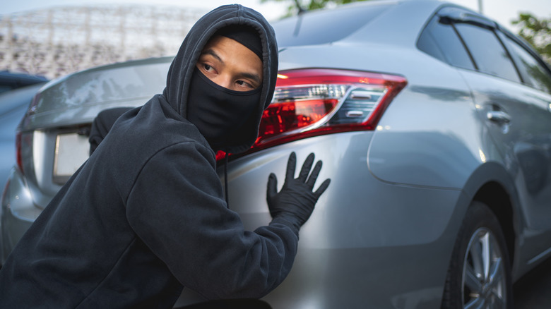 A Person Dressed In Black With Their Face Concealed Lurking Near A Parked Car