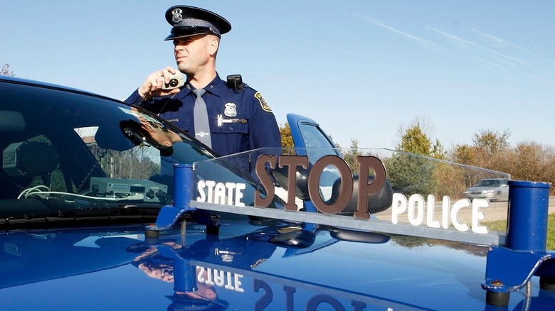 Michigan State Patrol officer standing next to his patrol car talking on the radio with Stop State Police Sign visible.