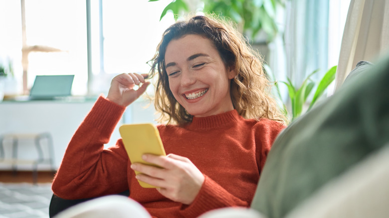 Happy relaxed young woman sitting on couch using cell phone