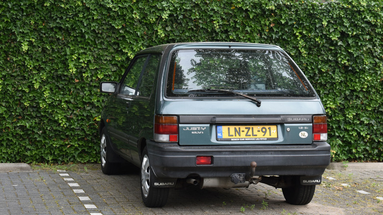 A green 1990 Subaru Justy parked against a vine-covered wall