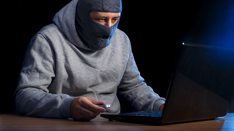 Man wearing balaclava holding USB drive next to laptop