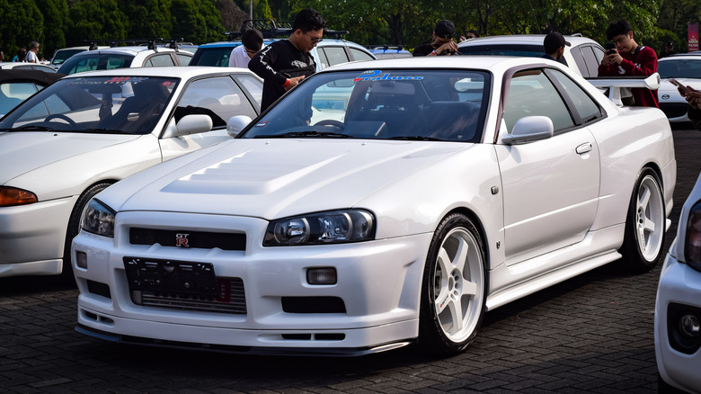 A modified R34 Nissan Skyline GT-R, white exterior with white wheels, front 3/4 view