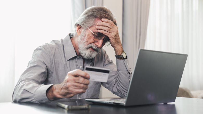 Elderly man dejectedly holding credit card in front of laptop