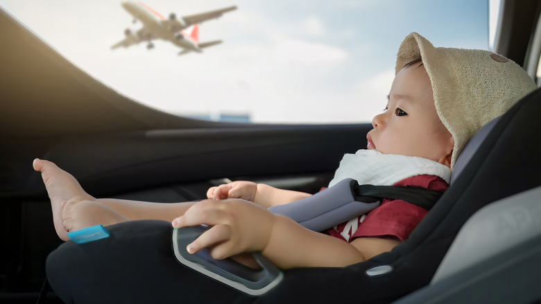 A baby is in a car seat while an airplane can seen through a window